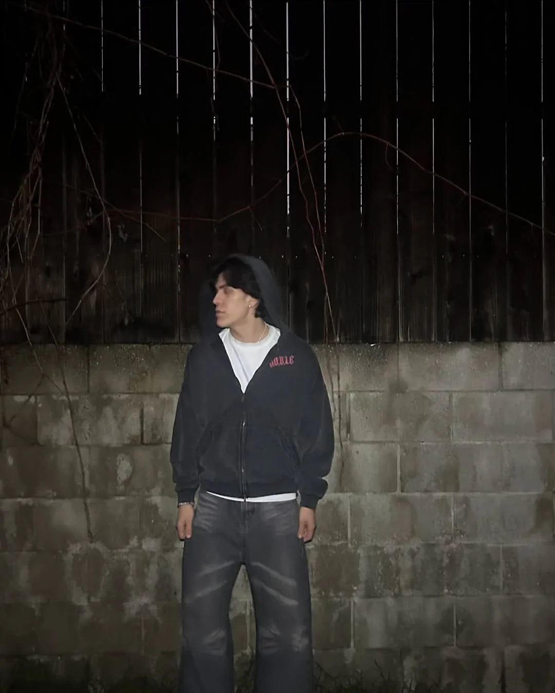 Person wearing a dark hoodie and jeans standing against a dark wall with a textured surface.
